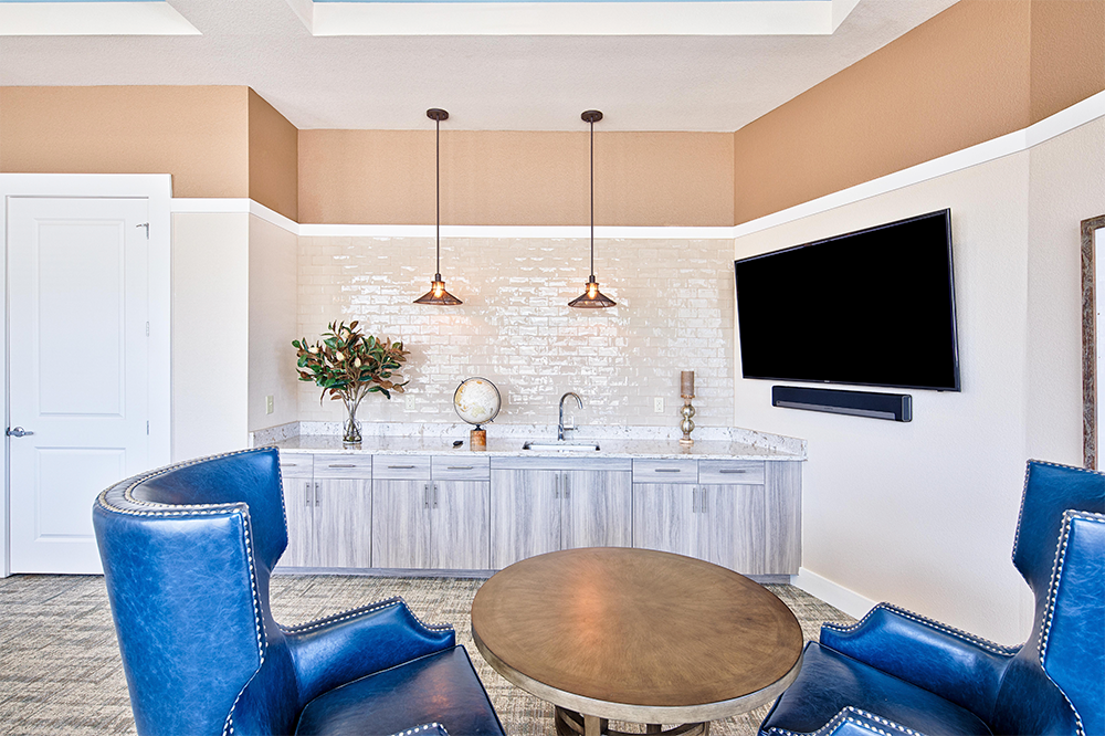 Modern lounge area with two blue armchairs, a round wooden table, wall-mounted TV, built-in cabinet, sink, and decorative items under pendant lights—perfectly suited for market rate developments and multifamily construction projects.