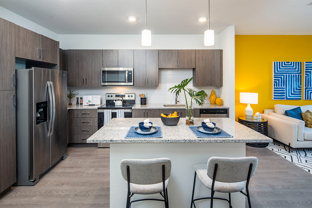 Modern kitchen with stainless steel appliances, gray cabinets, and a granite island with two chairs; perfect for market rate developments led by experienced multifamily general contractors. Living area features a yellow accent wall and blue artwork in the background.