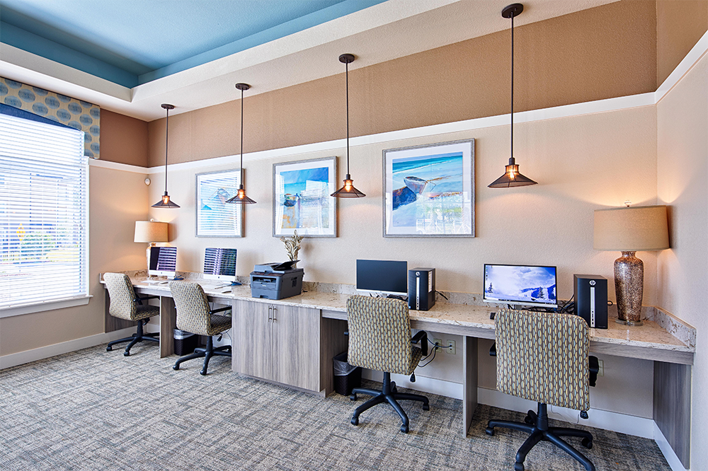 A modern office workspace with four desks, desktop computers, and a printer—ideal for multifamily general contractors or land development teams—features decorative lighting under framed art and a large window.