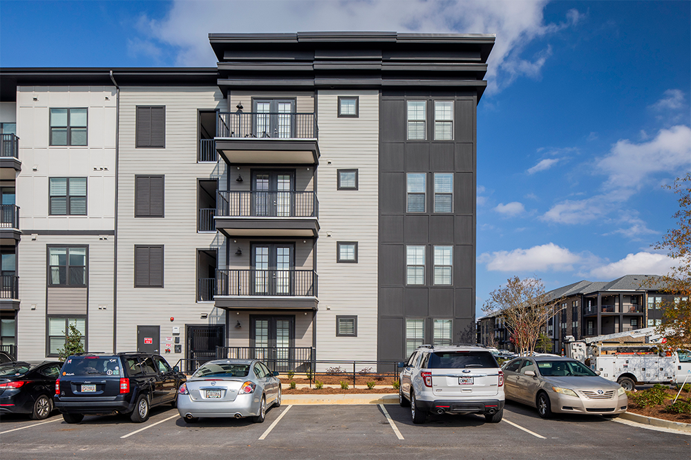 Four-story modern apartment building featuring balconies and gray siding, exemplifying contemporary multifamily construction. Several cars are parked in the lot out front beneath a partly cloudy sky.