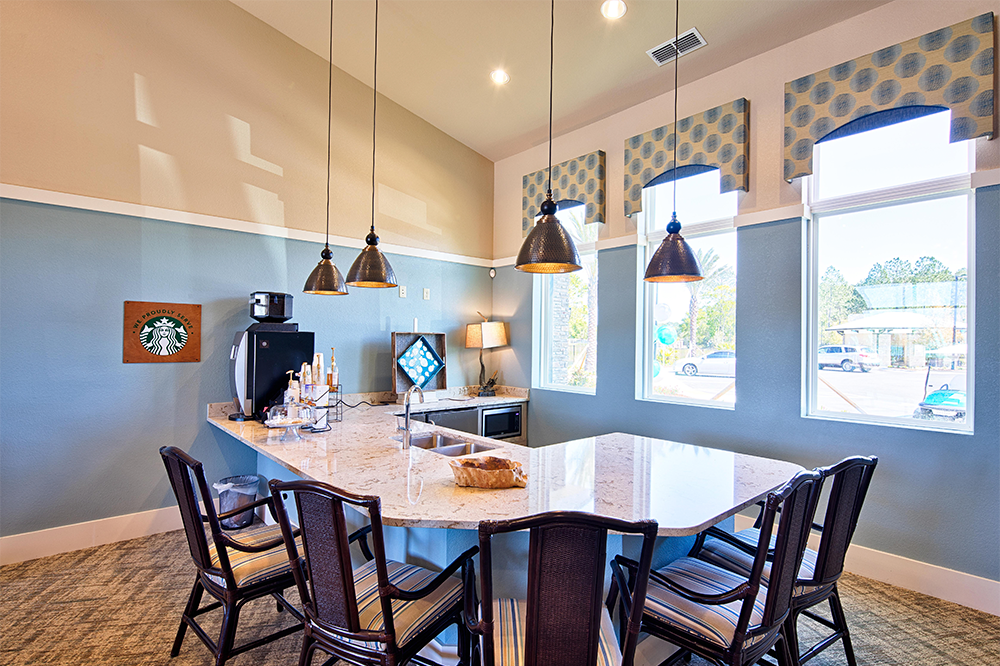 A modern kitchen with a large marble island, six chairs, pendant lights, coffee machine, and windows letting in natural light—perfect for market rate developments led by experienced multifamily general contractors.