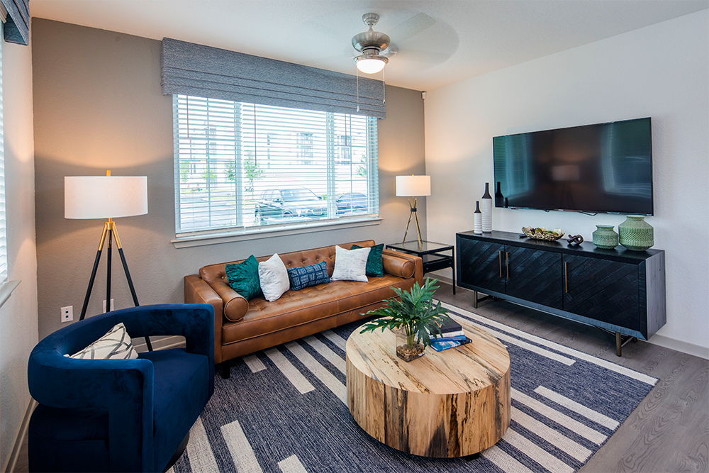 A modern living room, styled by multifamily general contractors, features a brown leather sofa, blue armchair, round wooden coffee table, striped rug, TV on the wall, and two floor lamps by a window with blinds.