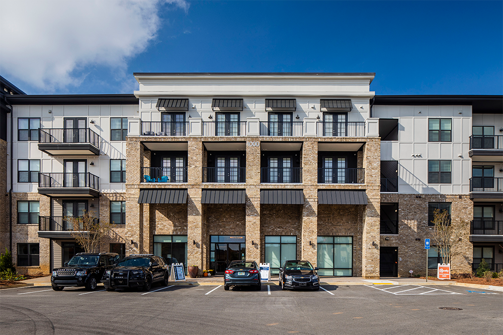 A modern three-story apartment building, showcasing market rate developments, features a brick and white panel exterior, balconies, and parked cars in front under a clear blue sky.