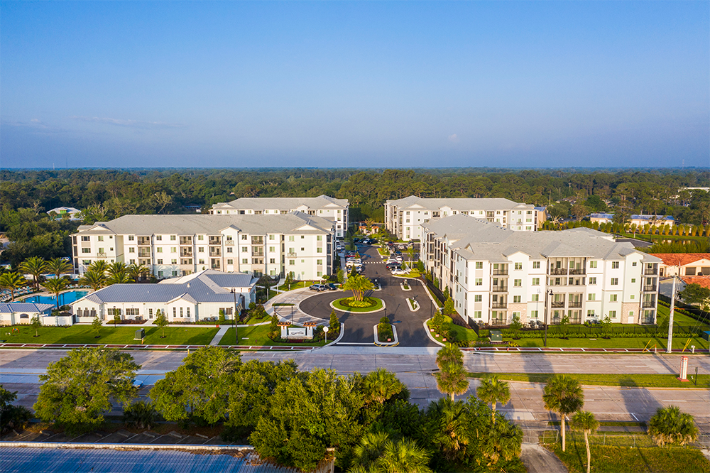 Aerial view of a modern apartment complex showcasing multifamily construction with white buildings, landscaped lawns, a central driveway, and surrounding trees under a clear blue sky.