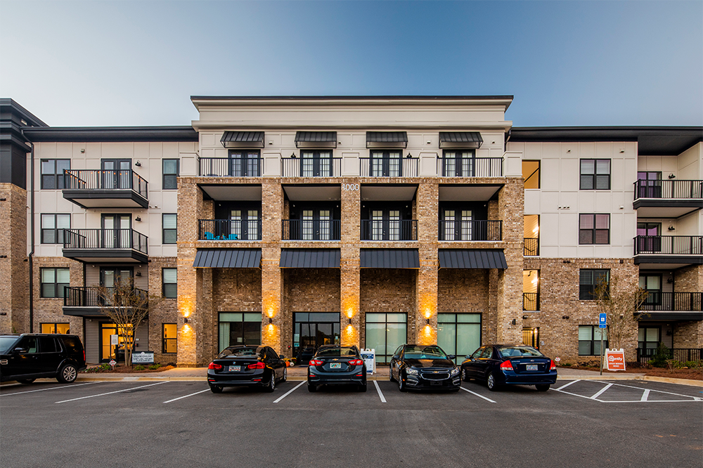 Four-story apartment building with brick and light-colored facade, balconies, and three parked cars in front of marked parking spaces—an excellent example of multifamily construction for market rate developments.