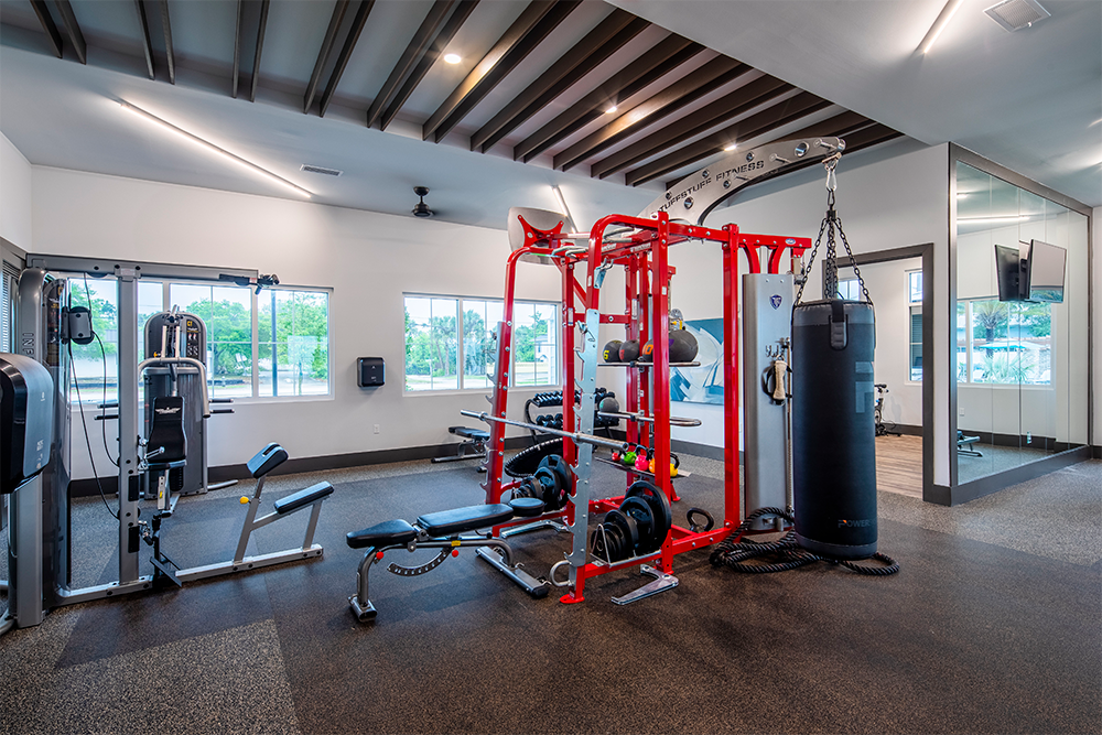 Modern gym room with weightlifting equipment, a punching bag, resistance machines, large windows, and a wall-mounted TV—expertly designed by multifamily general contractors for quality land development projects.