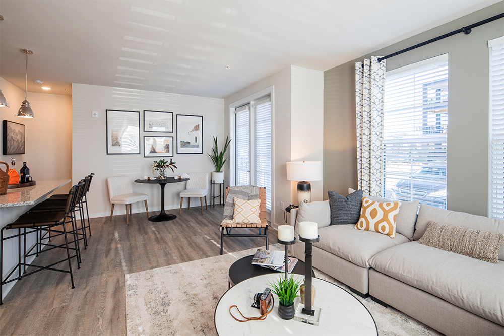 Modern open-concept living room and kitchen with neutral decor, gray sofa, round coffee table, dining area, and large windows letting in natural light—ideal for contemporary multifamily construction or upscale land development projects.