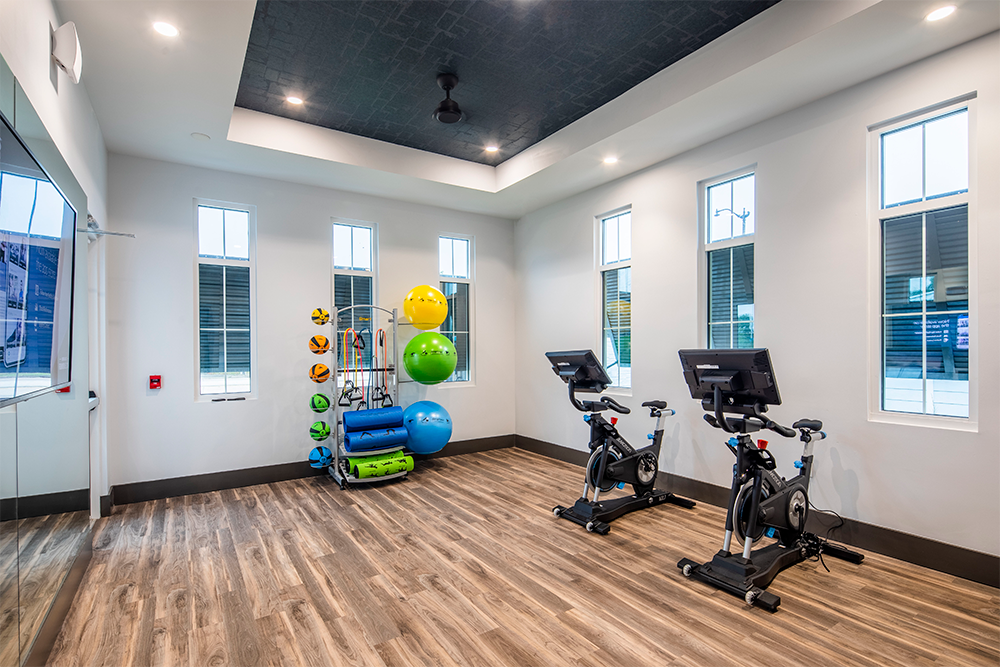 Modern fitness room designed for multifamily construction, featuring two stationary bikes, an exercise ball rack, yoga mats, and large windows that fill the space with natural light.