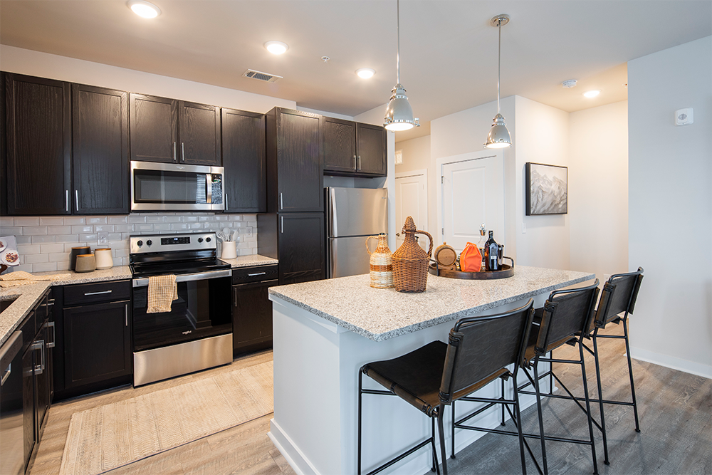 Modern kitchen with dark cabinets, stainless steel appliances, an island with granite countertop, three black barstools, and pendant lighting—perfect for multifamily general contractors designing refined market rate developments.