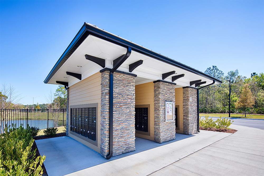 Outdoor community mailbox station with stone pillars and a metal roof, surrounded by greenery and pavement under a clear blue sky—ideal for land development or multifamily construction projects seeking both style and durability.