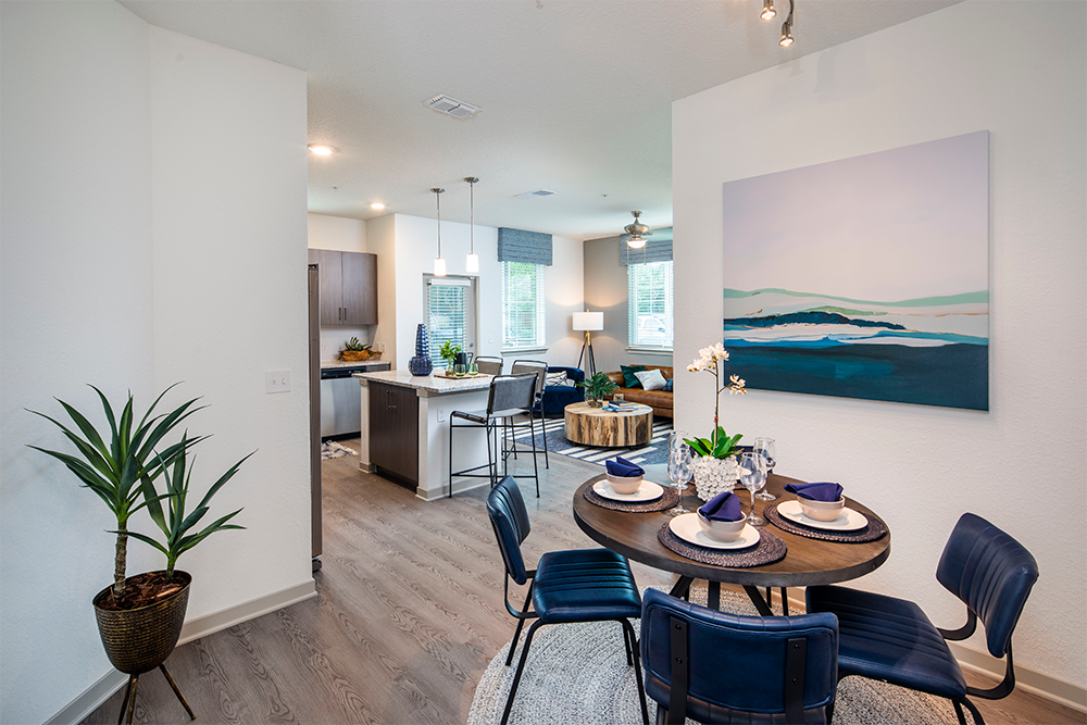 Modern open-concept apartment interior with a small dining area set for four, adjacent to a kitchen and living room featuring contemporary decor and large windows—an ideal example of quality multifamily construction.