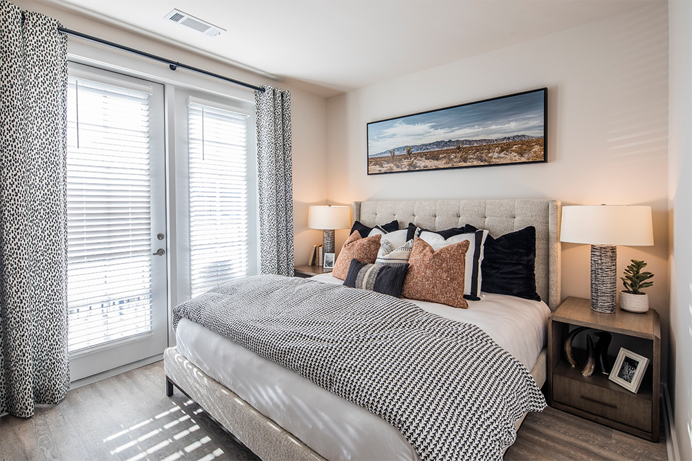 A modern bedroom designed for market rate developments, featuring a large bed with patterned bedding, two bedside tables with lamps, a plant, and a landscape photo above the headboard. Light enters through glass doors with blinds.