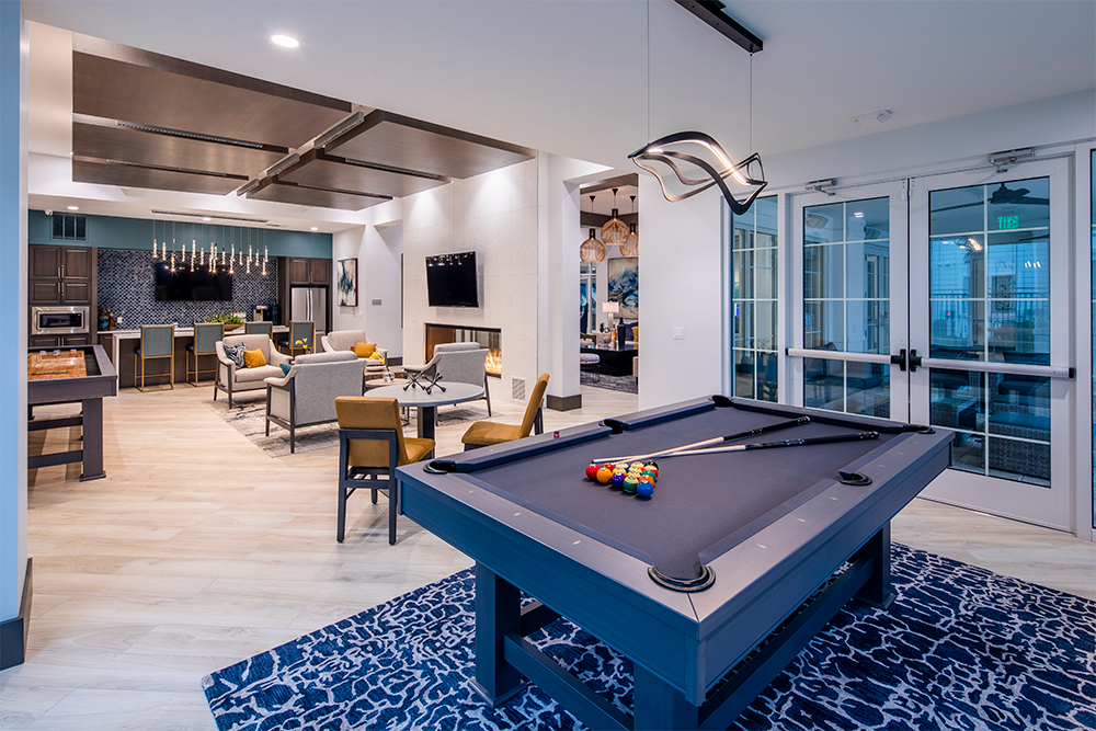 Modern lounge with a pool table in the foreground, seating area with TV, and open kitchen in the background; large windows on the right—an inviting space crafted by multifamily general contractors for contemporary living.