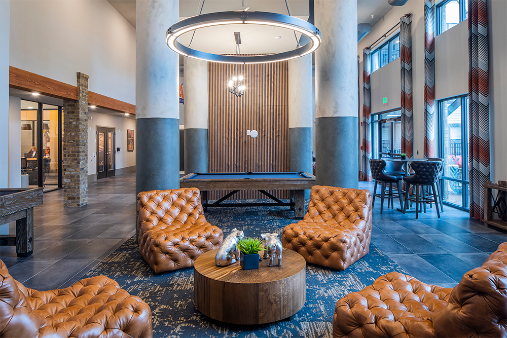 Modern lounge with tufted leather chairs, a round wooden coffee table, pool table, high ceilings, large windows, and industrial-style lighting—crafted by multifamily general contractors for upscale market rate developments.