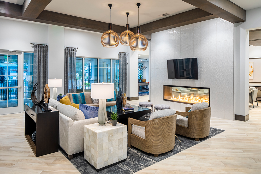 Modern living room with a sectional sofa, two armchairs, a wall-mounted TV, a double-sided fireplace, pendant lights, and large windows with curtains—ideal inspiration for multifamily construction or market rate developments.