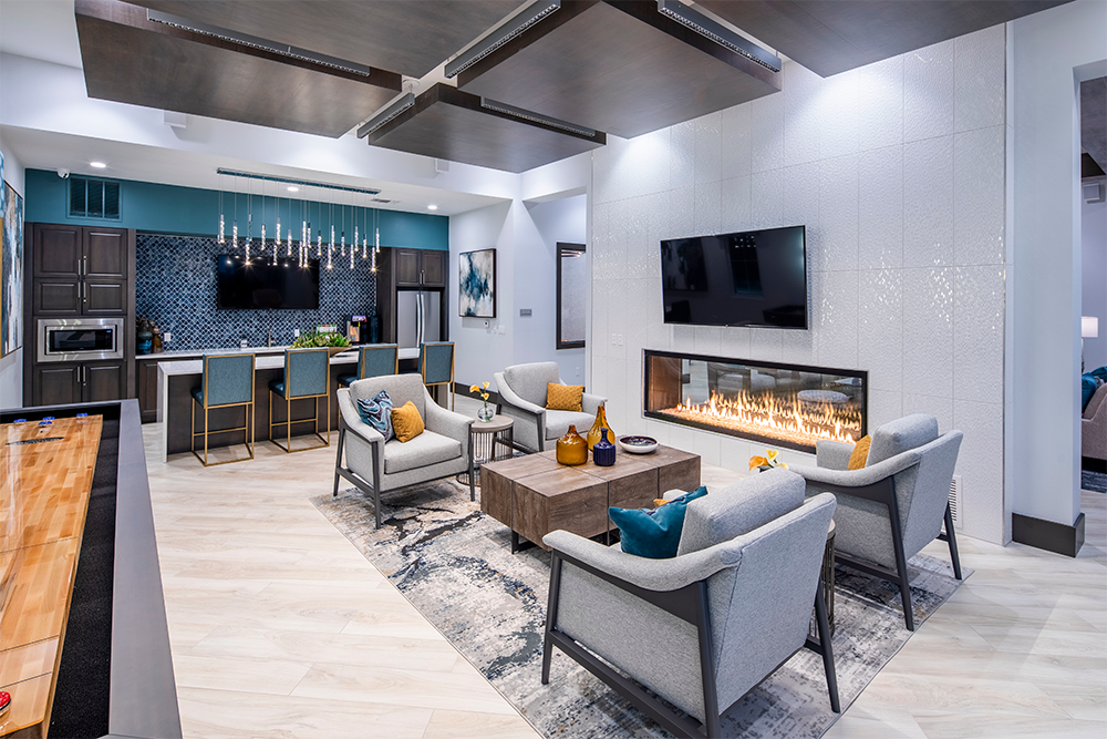 Modern living area with four gray armchairs around a wood coffee table, a wall-mounted TV above a linear fireplace, and an open kitchen with bar seating—a perfect showcase for multifamily construction and modern market rate developments.