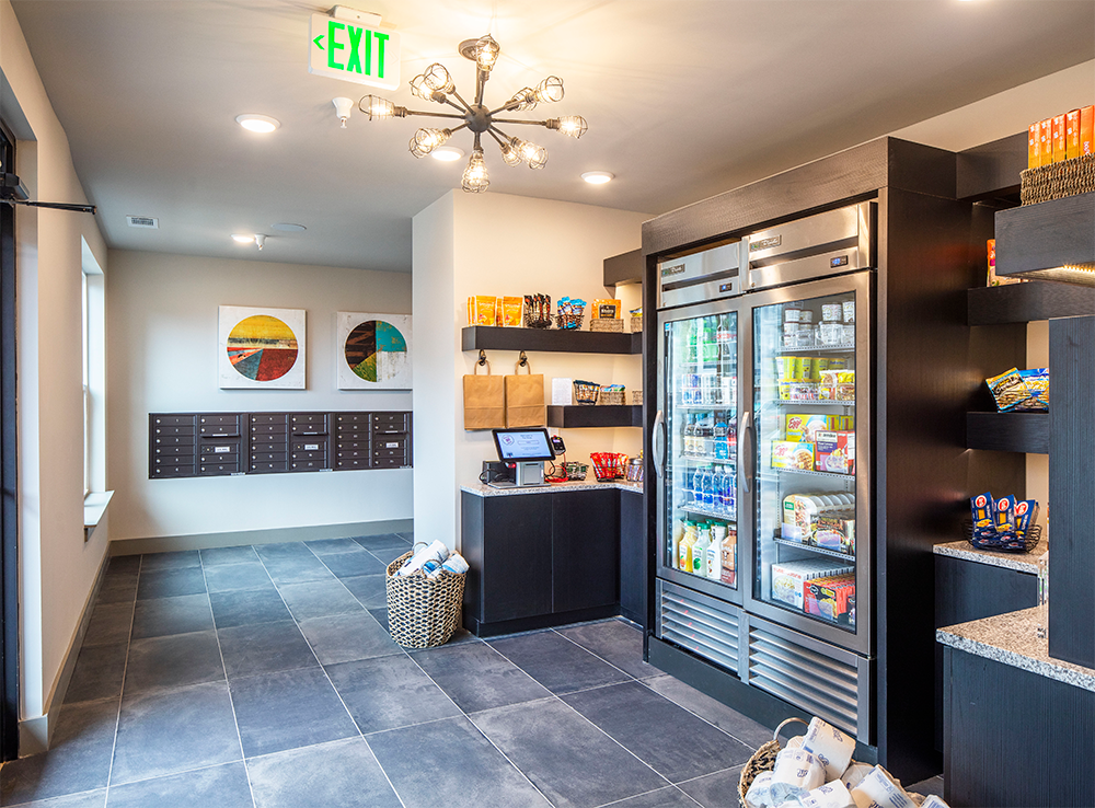 A small convenience area in market rate developments, featuring shelves and coolers stocked with snacks and drinks, a touchscreen kiosk, mailboxes, and modern decor under bright lighting.
