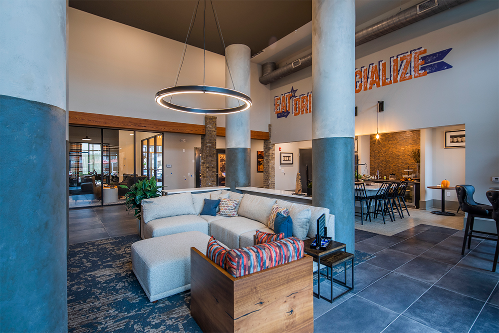 Modern lounge area with a large sectional sofa, circular ceiling light, tall columns, and open view to dining and workspaces in a loft-style interior—an inviting space perfect for market rate developments or multifamily general contractors.