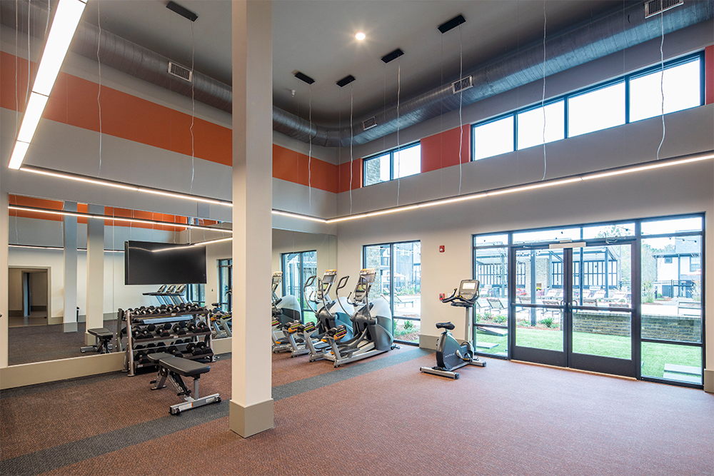 Modern gym with cardio machines, free weights, benches, large mirrors, and tall windows—expertly designed by multifamily general contractors—with a view of an outdoor patio area.