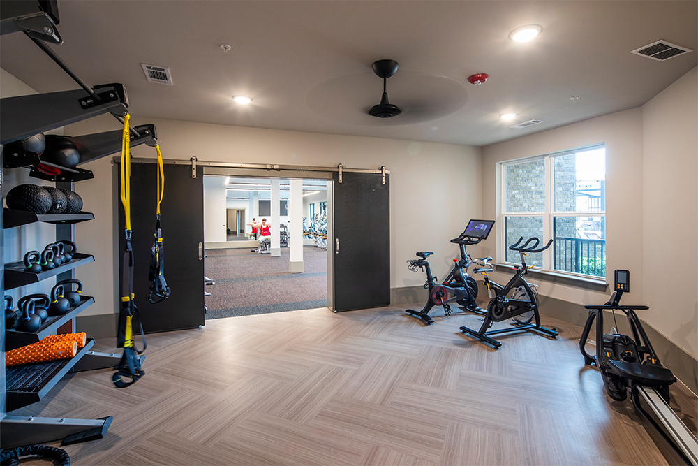 A small fitness room with exercise bikes, resistance bands, shelves with gym equipment, and an open doorway leading to a larger gym area—ideal for market rate developments and modern multifamily construction.