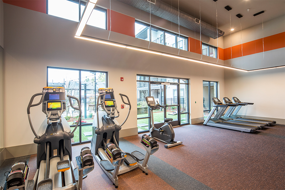 A modern gym room in market rate developments features three elliptical machines and three treadmills, large windows, high ceilings, and orange accents on the walls.