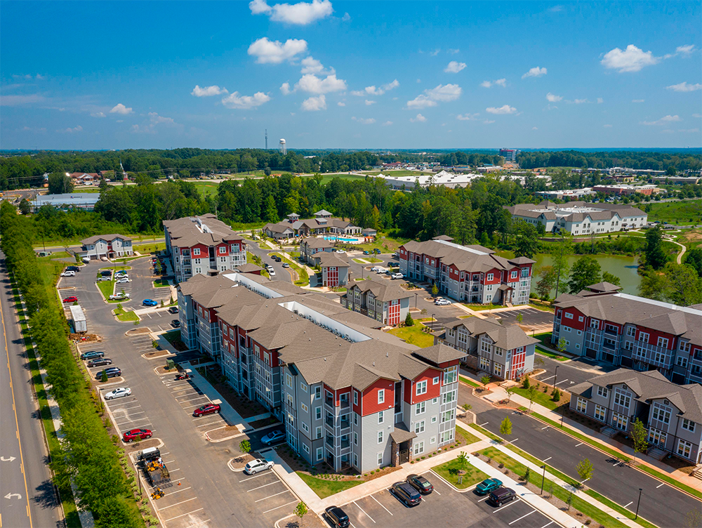 Aerial view of a modern apartment complex showcases multifamily construction with multiple buildings, parking lots, and surrounding greenery—an impressive example of market rate developments under a clear blue sky.