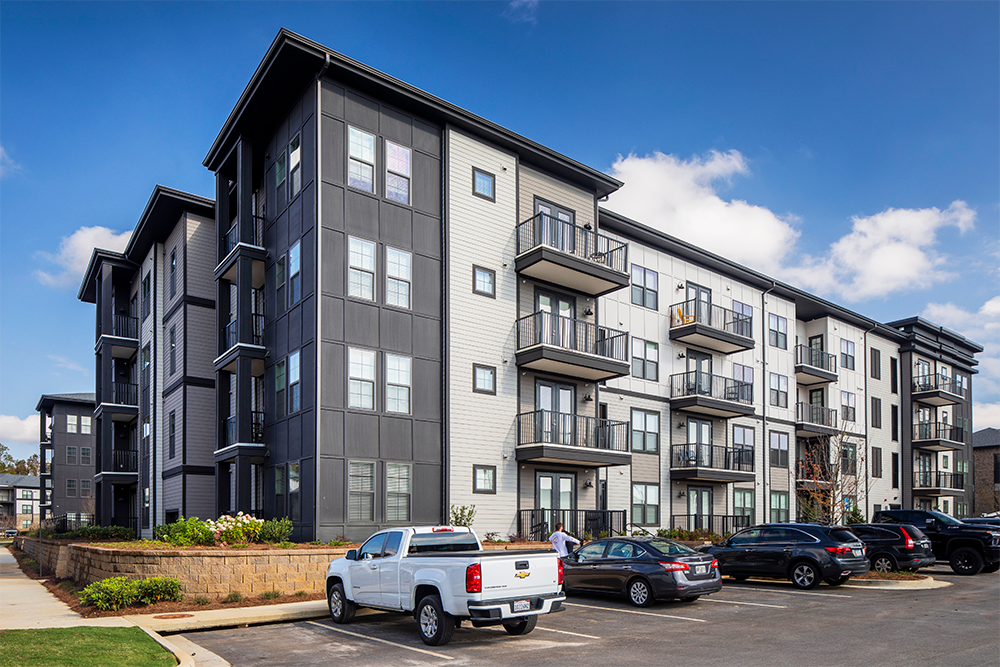 Four-story modern multifamily construction apartment building with balconies and parked cars in a lot on a clear day.