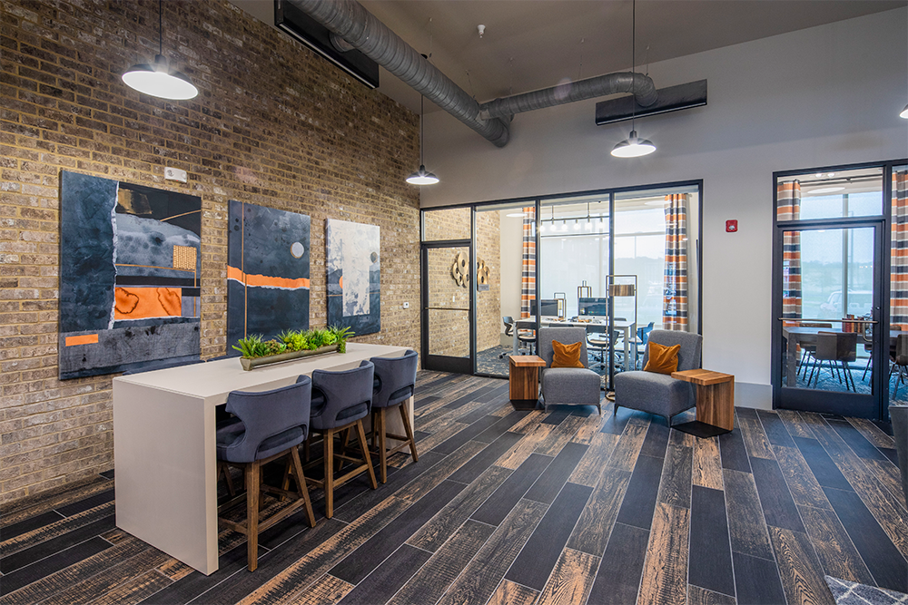 Modern office lounge with a high table and chairs, abstract wall art, large windows, and a meeting room visible through glass walls—designed by multifamily general contractors. Dark wood flooring and exposed brick walls complete the look.