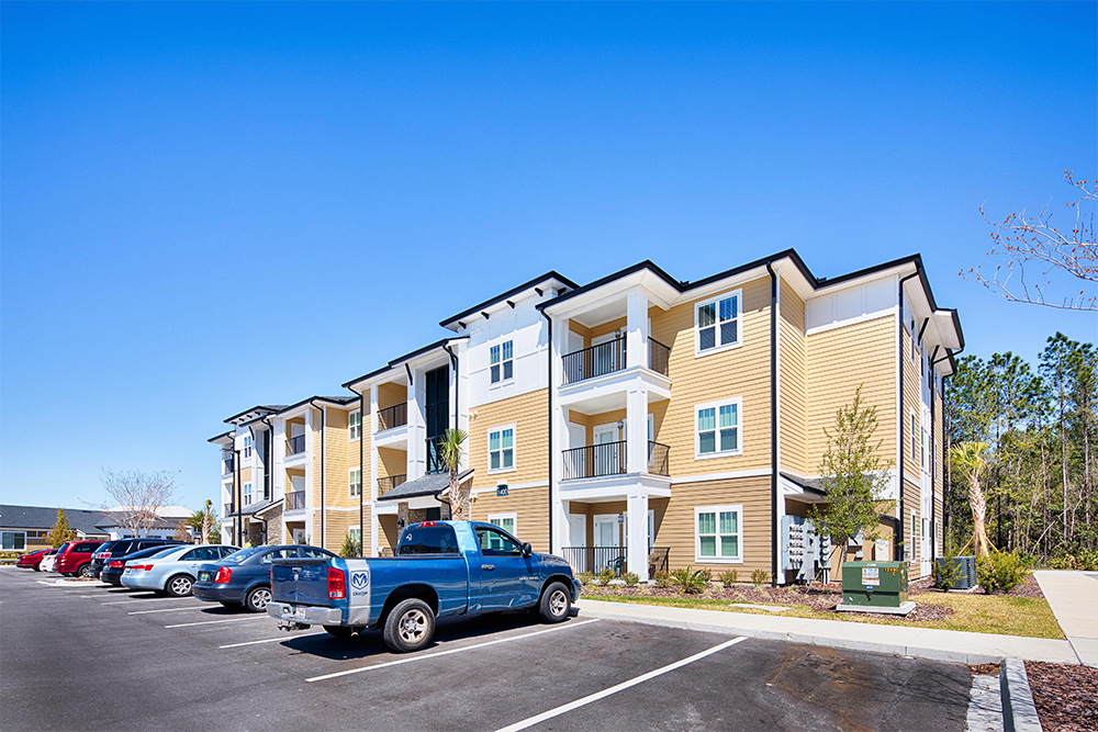 A three-story apartment building with balconies, yellow siding, and white trim stands next to a parking lot under a clear blue sky—an example of quality work by multifamily general contractors in market rate developments.