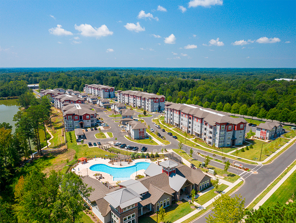 Aerial view of a modern apartment complex showcases multifamily construction with multiple buildings, a central swimming pool, parking lots, and surrounding trees under a clear blue sky.