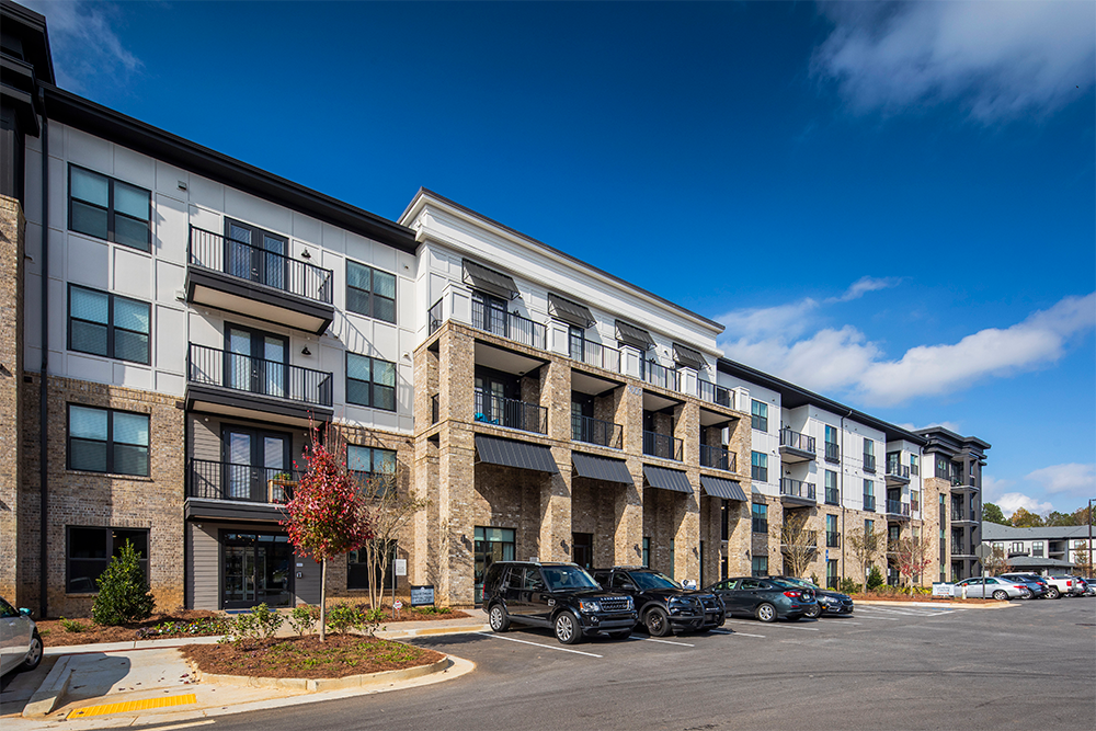 Three-story apartment building with balconies and brick exterior, constructed by expert multifamily general contractors, adjacent to a parking lot with several parked cars under a clear blue sky.