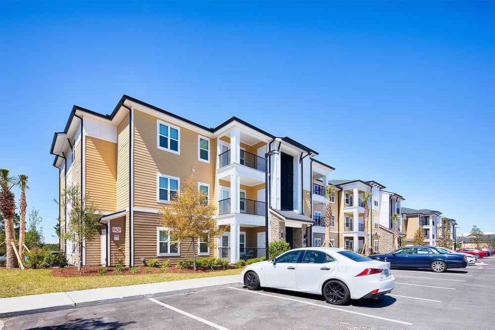 Three-story apartment building with beige siding and balconies, typical of market rate developments, viewed from a parking lot with a white car parked in front under a clear blue sky.