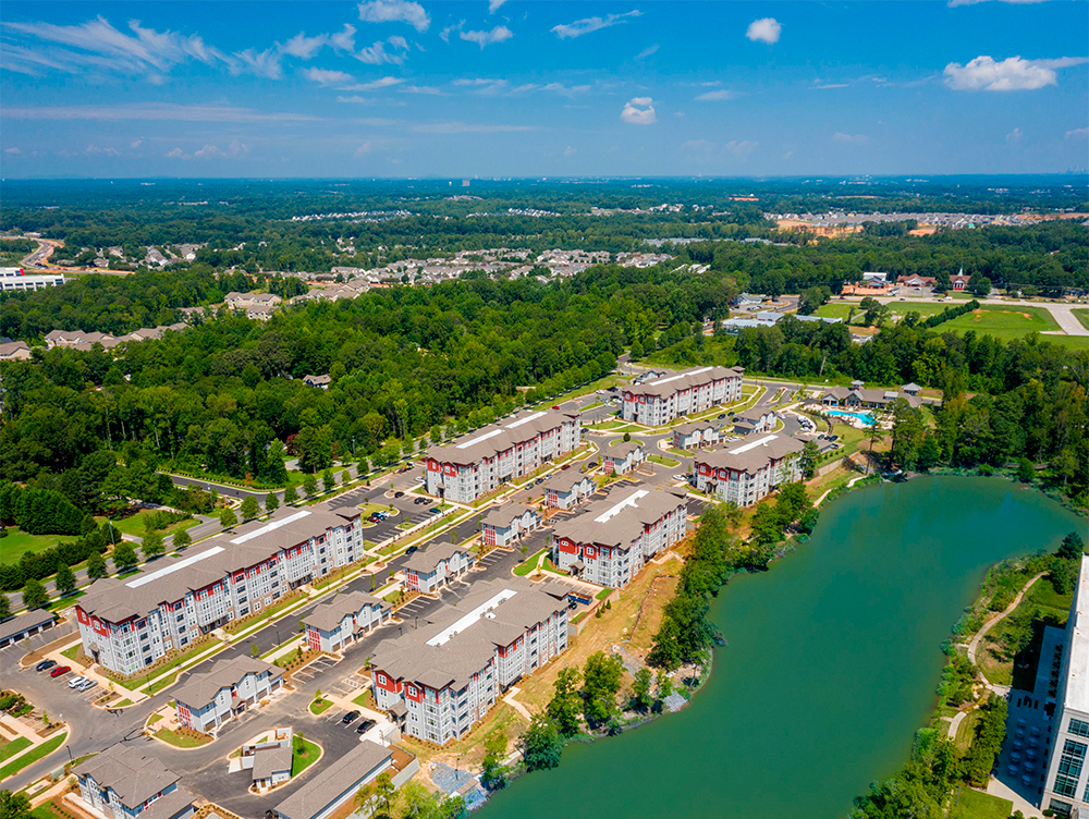 Aerial view of a residential apartment complex beside a lake, surrounded by trees, with a clear sky and suburban market rate developments in the background.