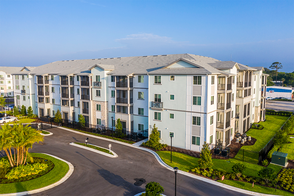 A four-story apartment building with light siding, balconies, and surrounding landscaping—an example of successful multifamily construction—viewed from above on a clear day.