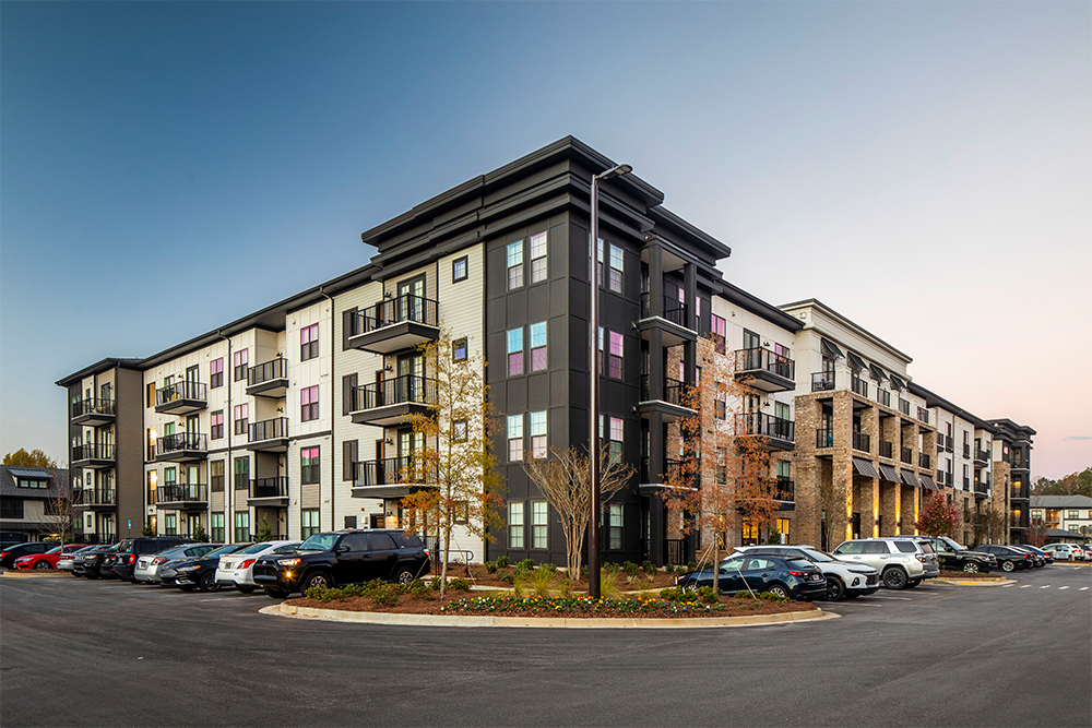 Modern four-story apartment building with balconies, surrounded by a parking lot filled with cars and landscaped trees in front, photographed at dusk—a prime example of market rate developments in contemporary land development.