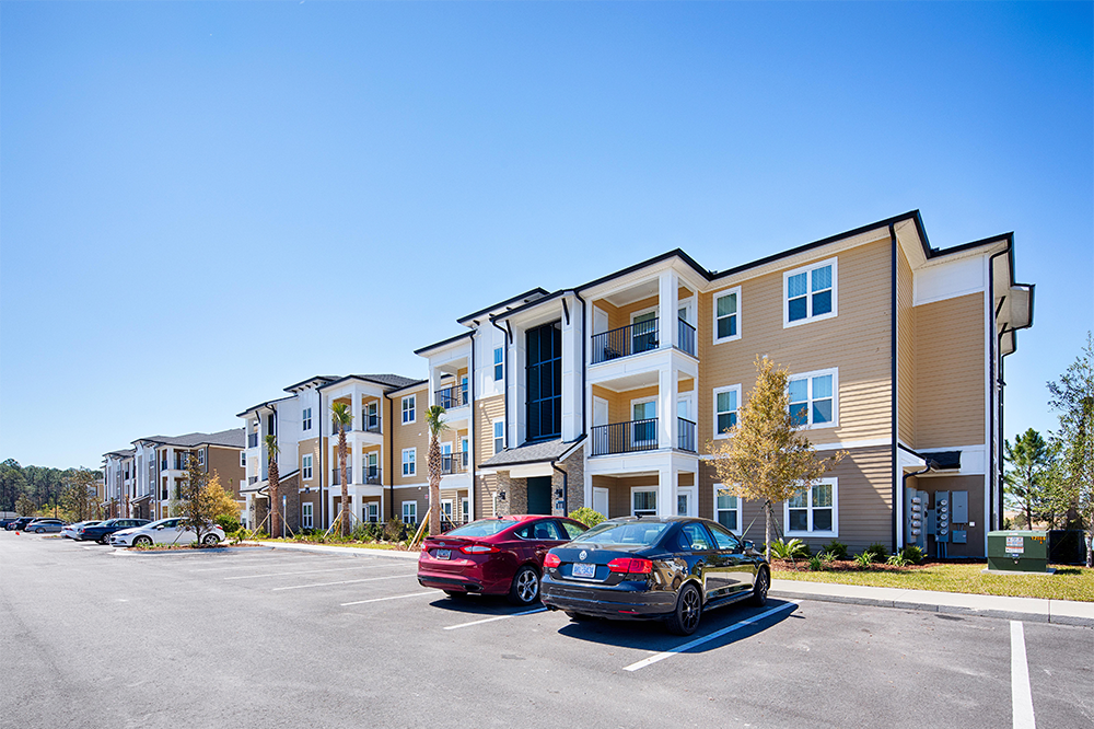 Three-story apartment buildings with balconies and parked cars in a paved lot under a clear blue sky, showcasing quality multifamily construction.