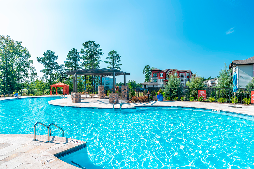 Outdoor swimming pool with clear blue water, surrounded by a stone deck, pergola, trees, and residential buildings—an inviting amenity commonly found in market rate developments built by leading multifamily general contractors.