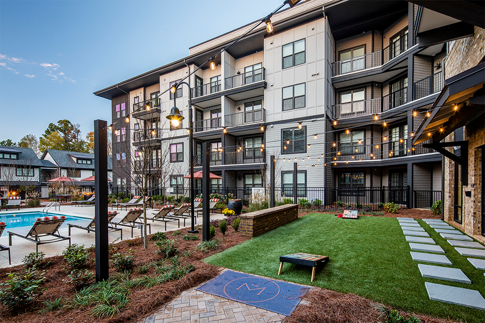 Modern multifamily construction with balconies overlooks a landscaped courtyard, swimming pool, lounge chairs, string lights, and a cornhole game setup on artificial turf.