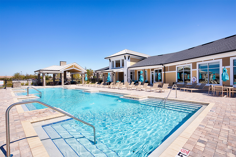 Outdoor swimming pool with lounge chairs and umbrellas beside a modern building under a clear blue sky, showcasing the quality expected from leading multifamily general contractors in market rate developments.