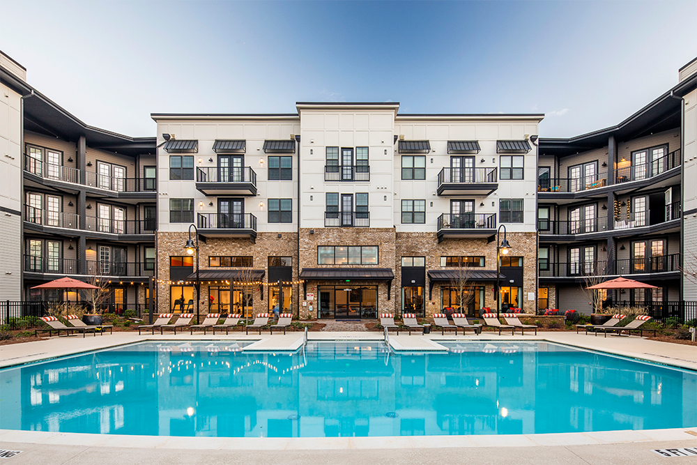 Modern apartment building with balconies and large windows, showcasing expert multifamily construction, faces a rectangular outdoor swimming pool surrounded by lounge chairs and umbrellas.