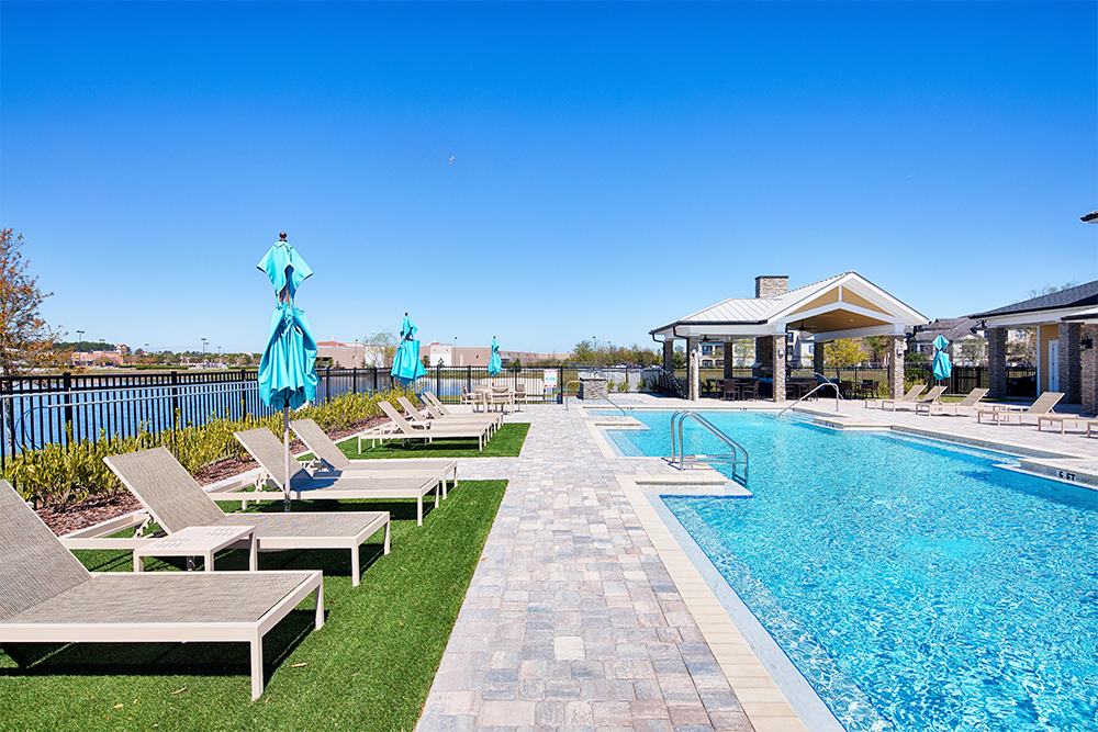 Outdoor swimming pool with lounge chairs, closed blue umbrellas, and a covered patio area, set beside a lake on a clear sunny day—ideal for market rate developments by multifamily general contractors.