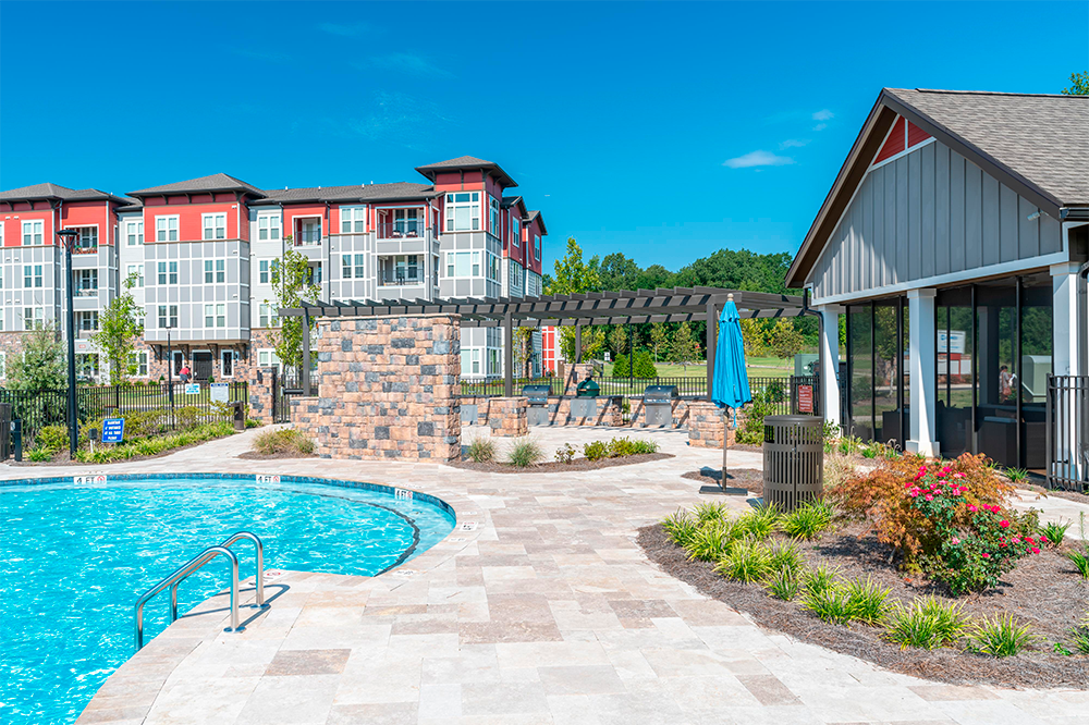 Outdoor swimming pool with curved edge, stone patio, lounge area, and surrounding apartment buildings under a clear blue sky—crafted by expert multifamily general contractors.