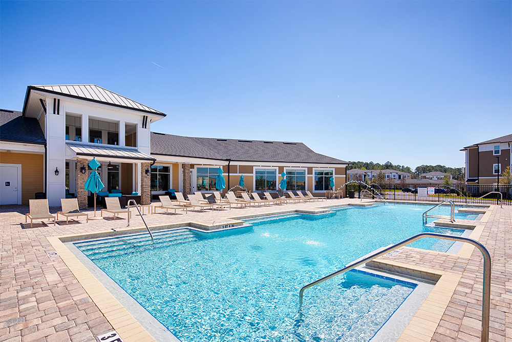 Outdoor swimming pool with lounge chairs and umbrellas, adjacent to a modern multifamily construction building under a clear blue sky.