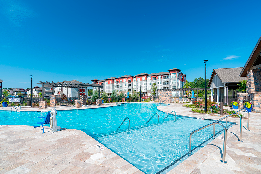 Outdoor swimming pool with clear blue water, surrounded by lounge chairs and buildings with red and white accents under a clear blue sky—perfect for modern market rate developments or multifamily construction projects.