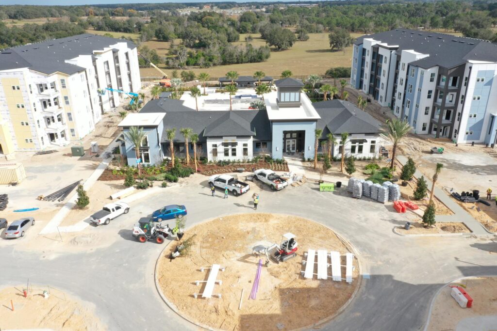 Aerial view of a multifamily construction apartment complex under development, with workers, vehicles, landscaping materials, and unfinished roadways visible around the central roundabout and main building.