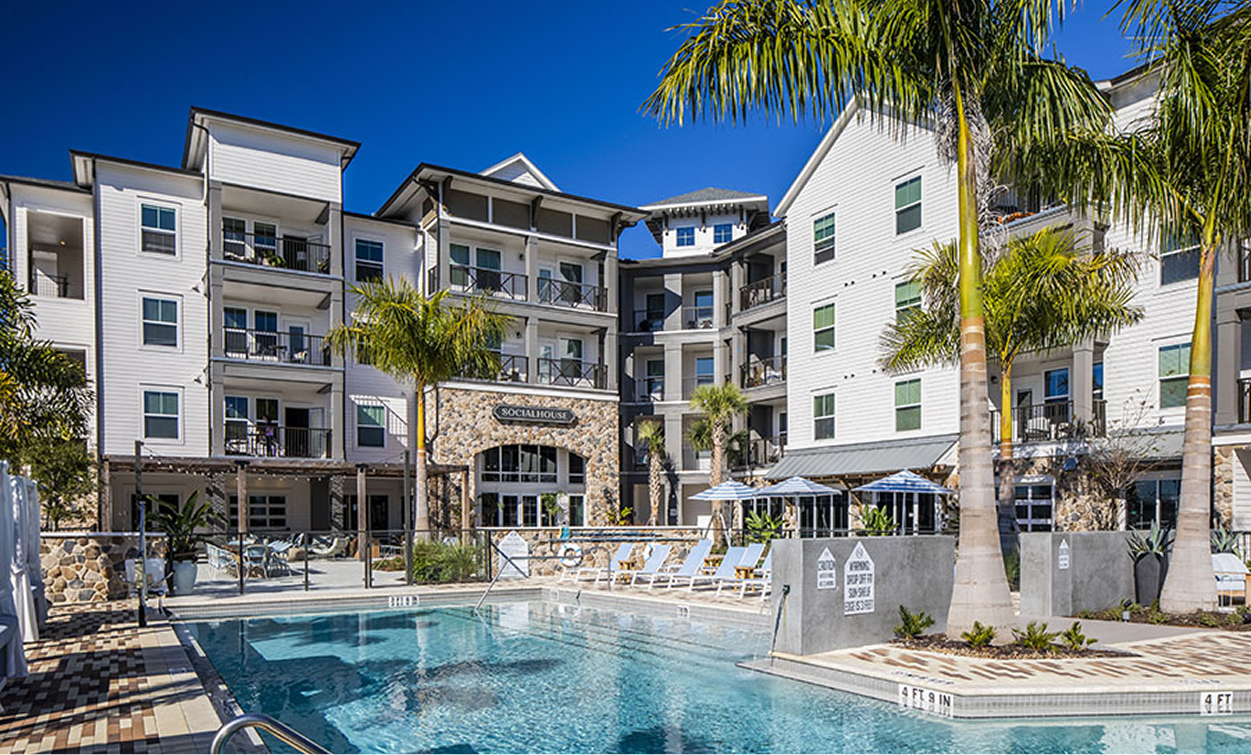 Modern apartment complex built by multifamily general contractors, featuring balconies overlooking a swimming pool surrounded by palm trees and lounge chairs on a sunny day.