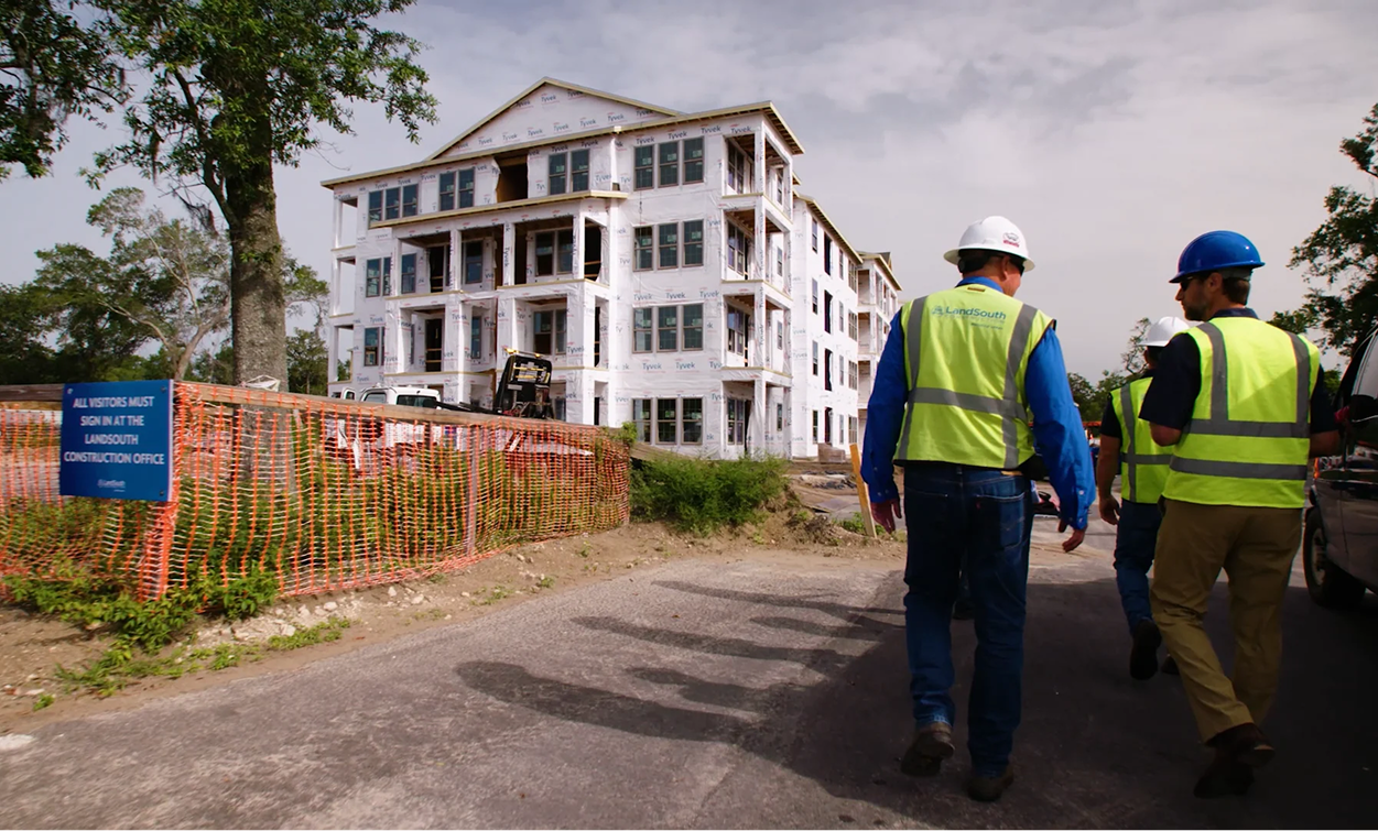 Construction workers in safety vests and helmets walk near a partially built, multi-story building wrapped in white house wrap at a fenced-off multifamily construction site.