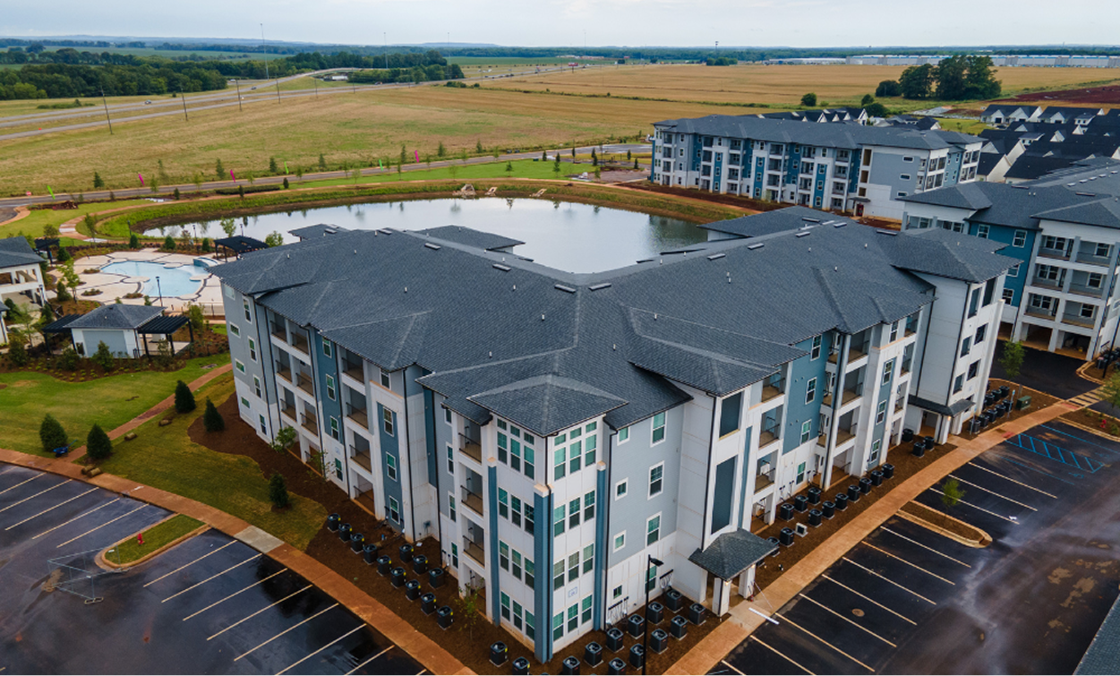 Aerial view of a modern multifamily construction apartment complex with four-story buildings, a pond, landscaped grounds, and adjacent parking lots. Fields and a highway are visible in the background.