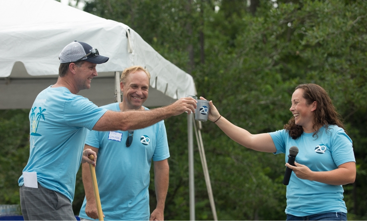 Three people in matching blue shirts celebrate outdoors; two men toast mugs while a woman with a microphone smiles and holds out her mug—an energetic moment at a multifamily construction event, with a white tent and trees in the background.