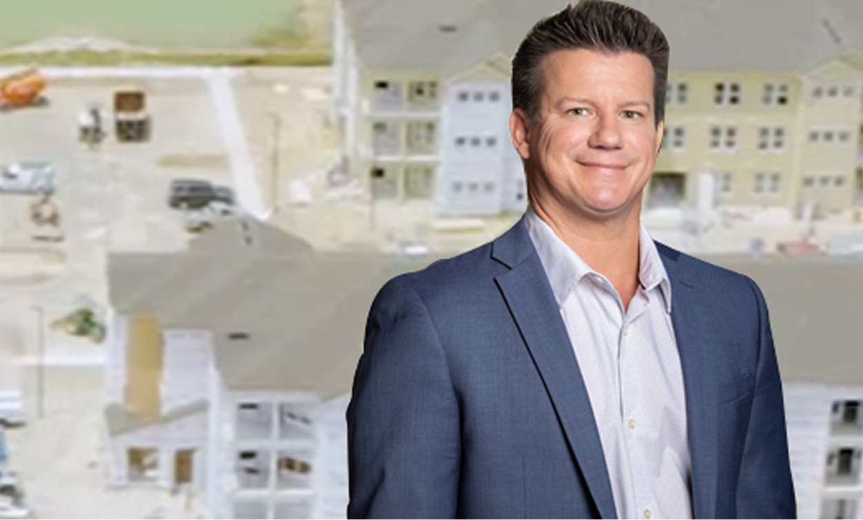 A man in a blue suit jacket and white shirt stands in front of a blurred background showing multifamily construction and buildings under development.